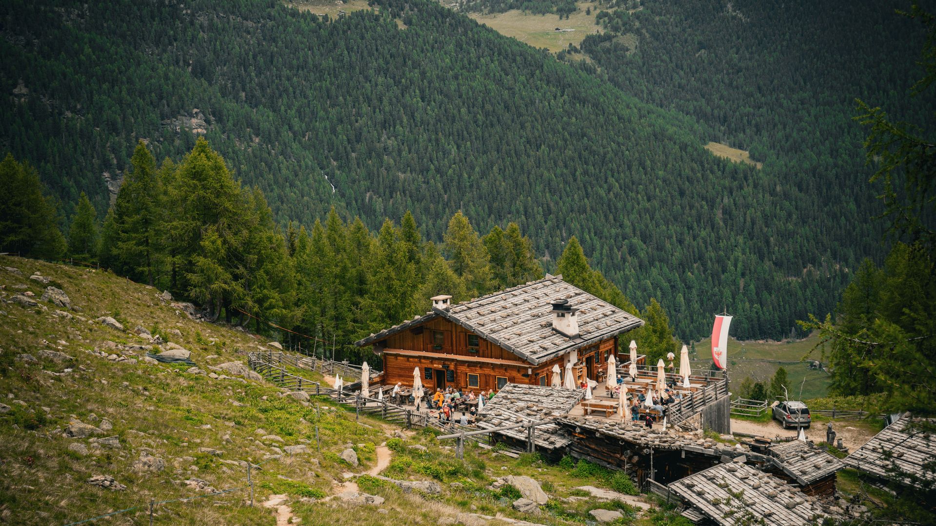 Almhütte mit Sonnenterrasse und Steindach an einem Bergwiesenhang, dahinter ein bewaldeter Talkessel