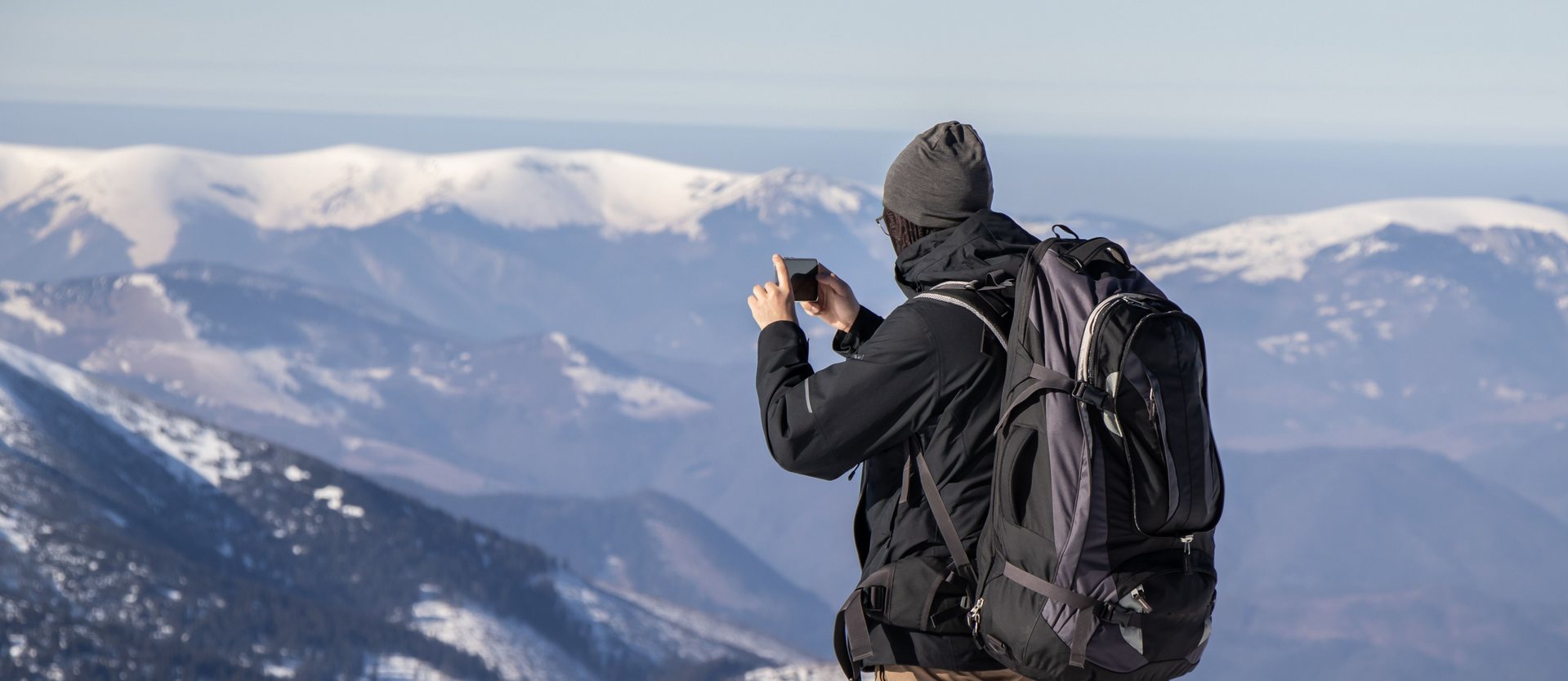 Person mit großem Rucksack fotografiert von einem Grat aus eine Kette verschneiter Bergrücken